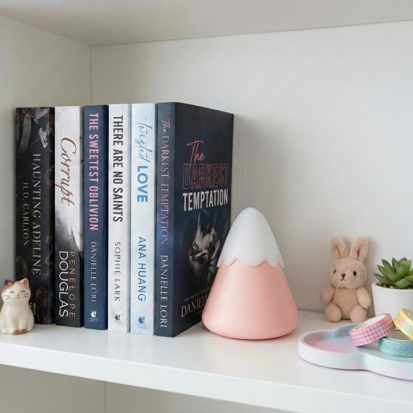 Pink Pika Fuji volcano clitoral suction sex toy on bookshelf with romance novels, cat figurine, bunny plush, succulent plant