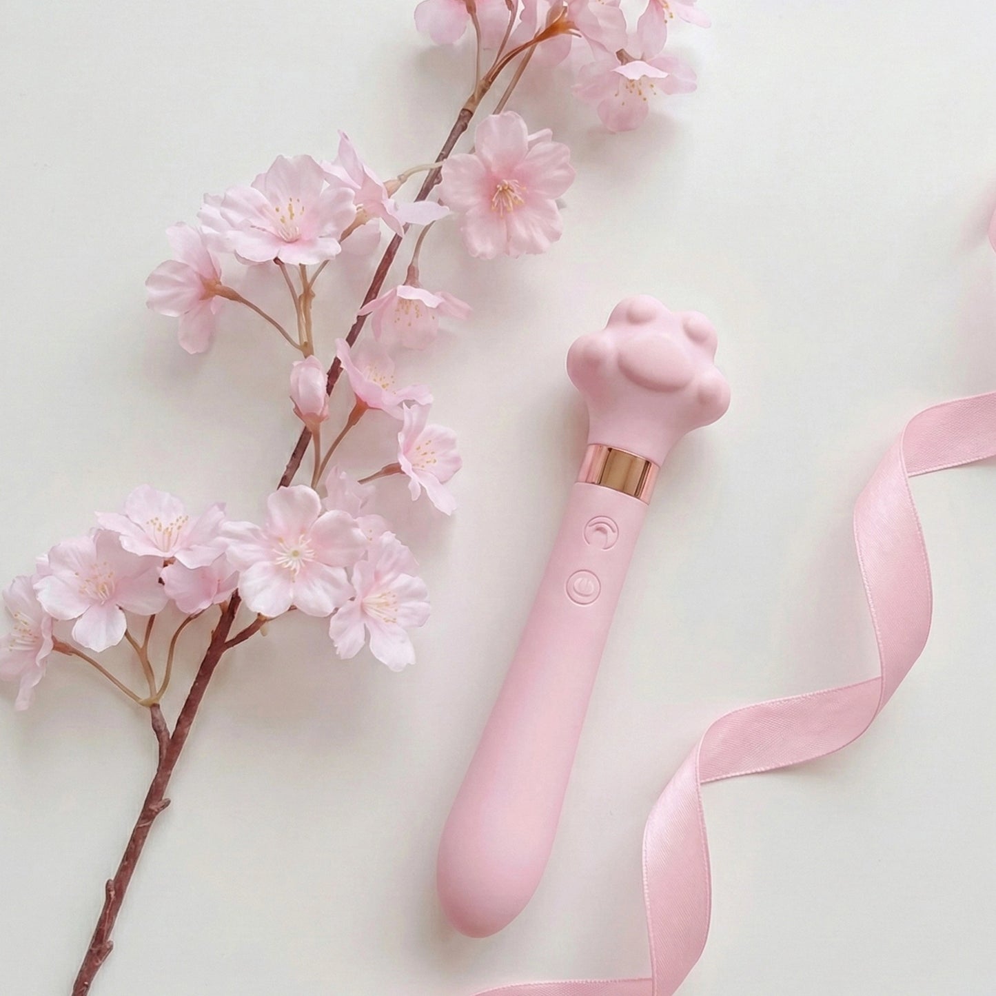 Pink cat paw-shaped wand vibrator with rose gold accent band displayed on white surface alongside pink cherry blossom branch and pink satin ribbon