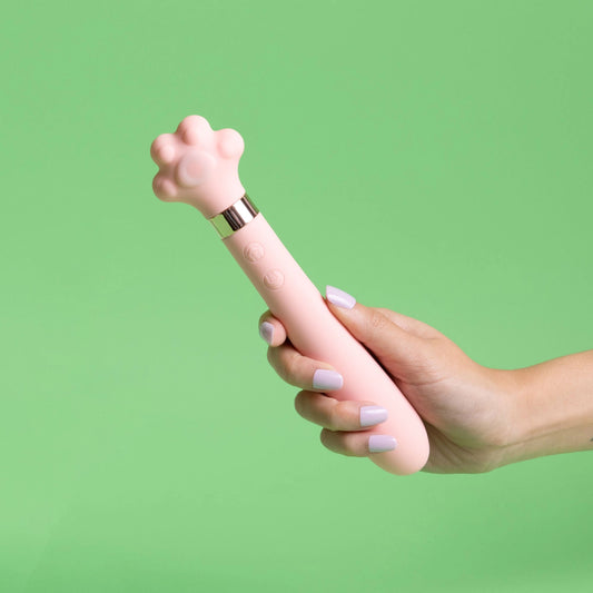 Hand with lavender manicure holding pink cat paw-shaped wand vibrator with rose gold metallic band against mint green background