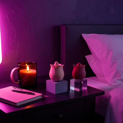 Two tulip-shaped clit licking vibrators, one pale pink and one dark red, displayed on a dark bedside table next to a lit candle, notebook, and bed in a dimly lit bedroom under purple ambient lighting.