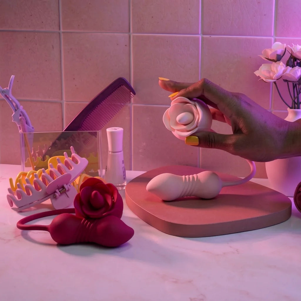 A hand holds a small pink rose toy personal massager over a pink tray, with additional pink and white intimate accessories arranged on a countertop against a tiled wall under pink lighting.