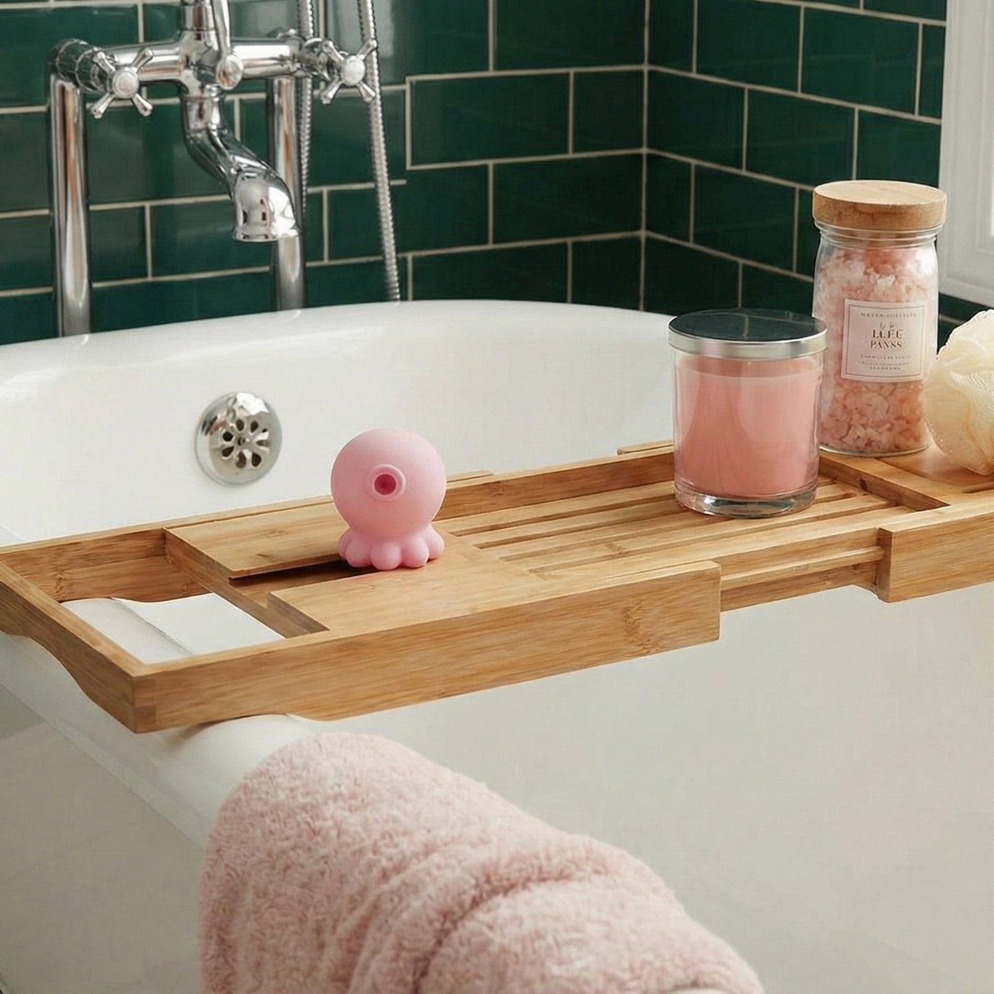 Pink octopus-shaped suction toy on bamboo bath caddy across bathtub, displayed with pink candle, bath salts jar, green tile wall, and fluffy pink towel