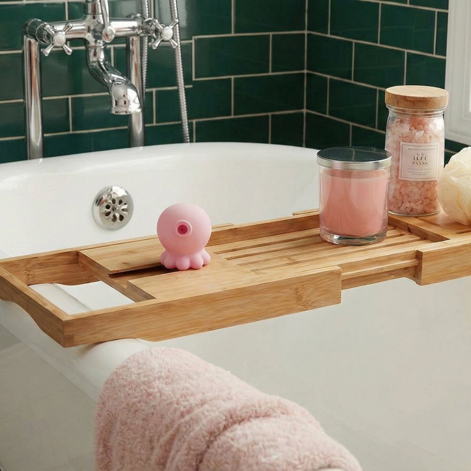 Pink octopus-shaped suction toy on bamboo bath caddy across bathtub, displayed with pink candle, bath salts jar, green tile wall, and fluffy pink towel
