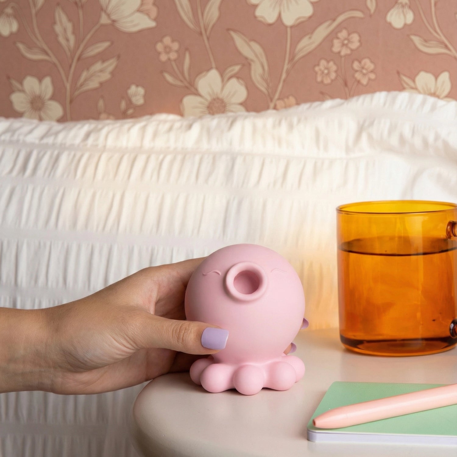 Hand holding pink octopus-shaped suction toy on a nightstand, shown with amber glass tumbler, white cable-knit textile in background, and pink and mint accessories
