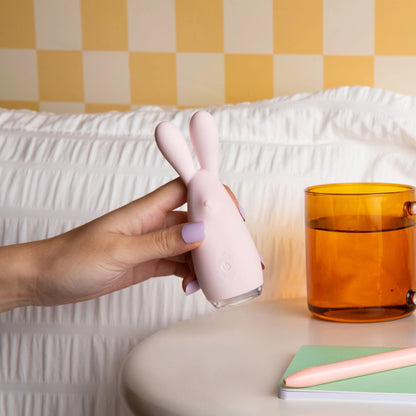 Hand with lavender nails holding pink bunny vibrator with LED light, white cable-knit background, amber glass tumbler