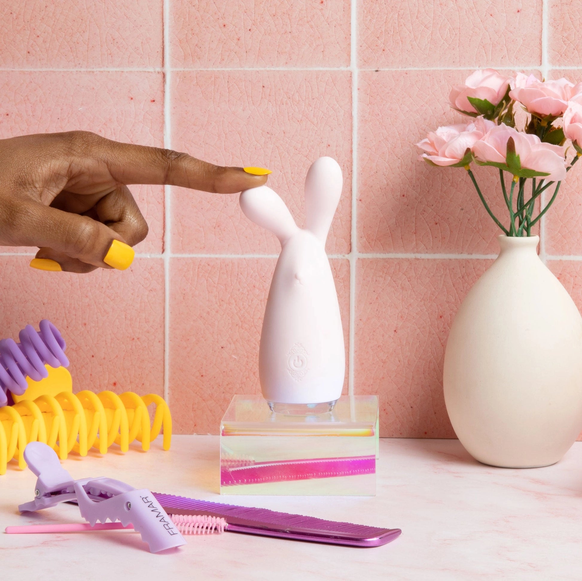 White bunny rabbit vibrator on acrylic stand with hand, pink bathroom tiles, roses in vase, and purple hair accessories