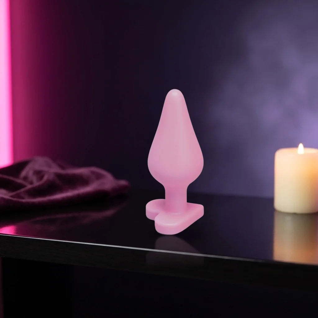Pink heart-base silicone plug standing on a dark shelf by candlelight