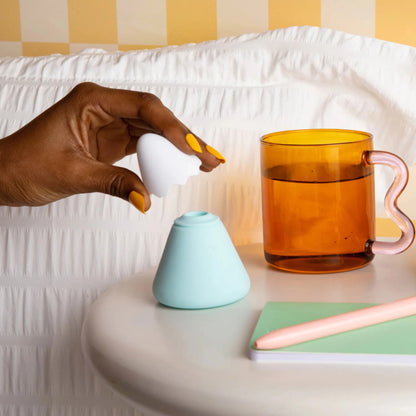 Hand with yellow manicure touching mint turquoise Fuji mountain clit sucker toy on nightstand with amber mug, gingham wallpaper