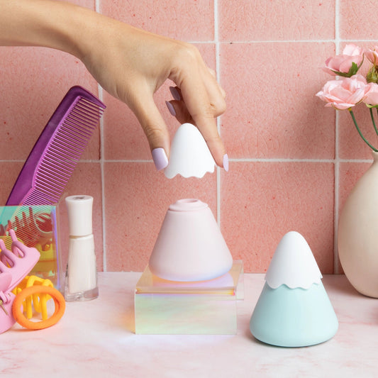 Hand with lavender nails holding Pika Fuji mountain clitoral suction sex toys in pink and mint on acrylic stand, pink tile, roses