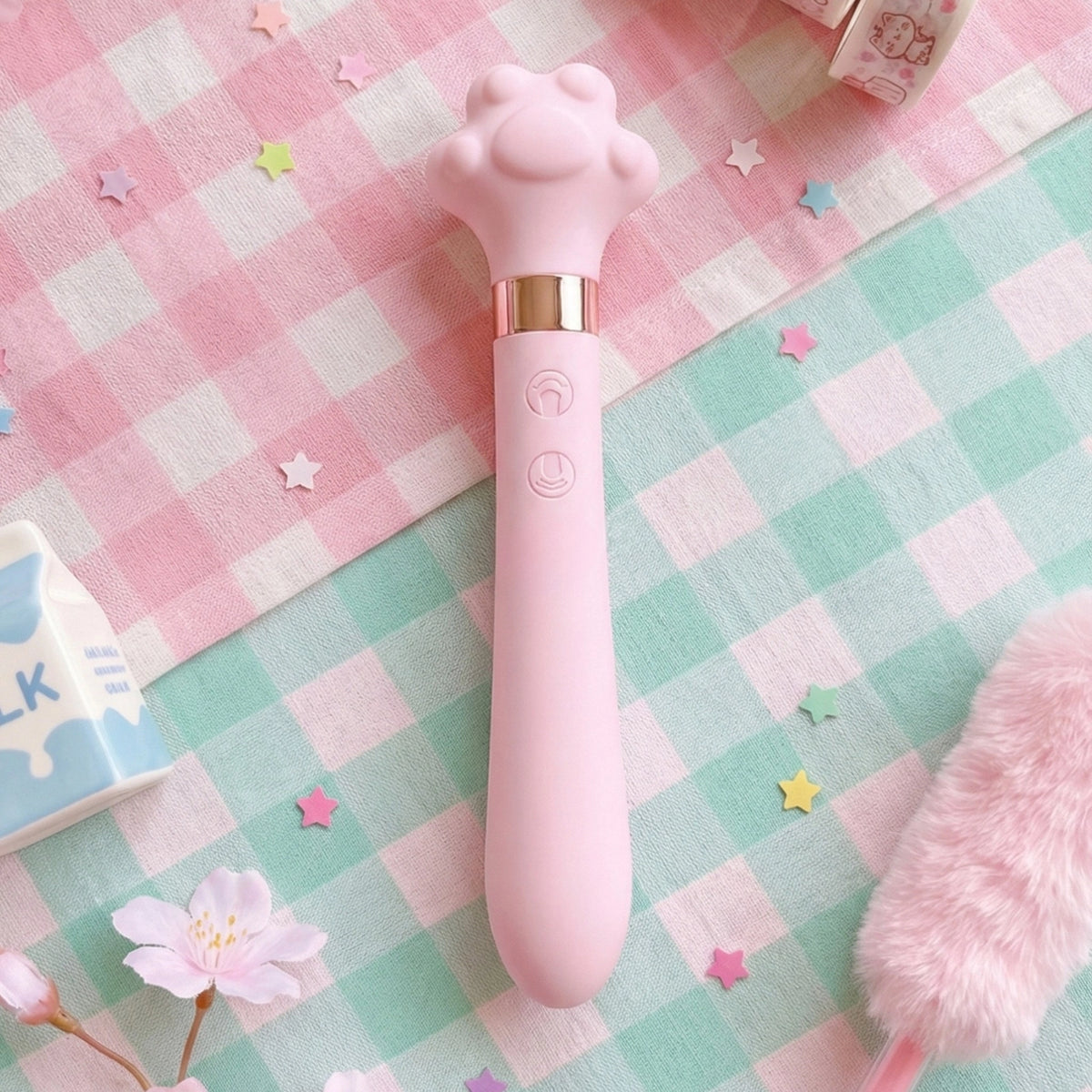 Pink cat paw-shaped wand vibrator on pastel pink and mint checkered blanket, surrounded by colorful star confetti, cherry blossom, pink fur accessory, and milk carton in background