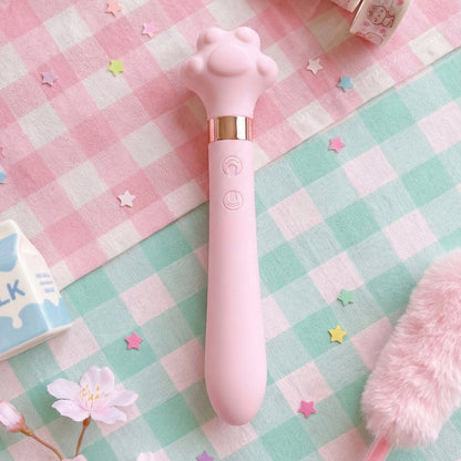Pink cat paw-shaped wand vibrator on pastel pink and mint checkered blanket, surrounded by colorful star confetti, cherry blossom, pink fur accessory, and milk carton in background