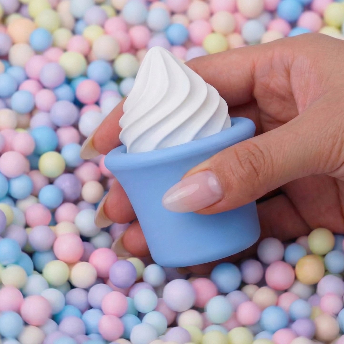 Hands holding blue Sprinkles soft serve cone clitoral vibrator sex toy against pastel candy ball pit background