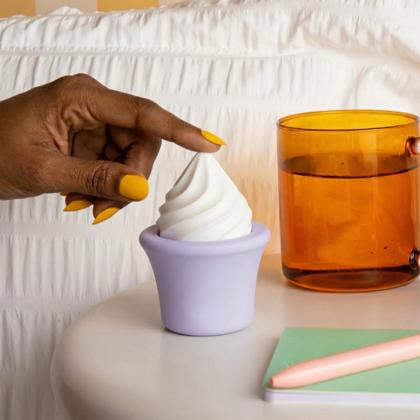Hand with yellow manicure touching lavender Pika Sprinkles ice cream cone vibrator on nightstand with amber glass, white bedding