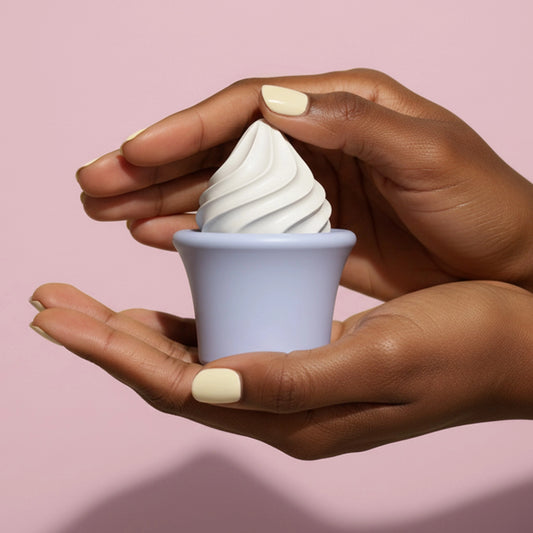 Hands with yellow nails cradling lavender ice cream cone shaped Pika vibrator against pink background
